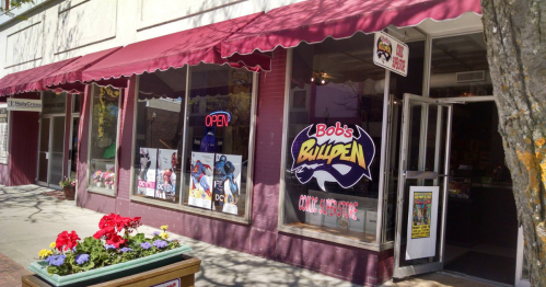 Exterior of Bob's Bullpen comic book store, featuring colorful displays and an "OPEN" sign, with flowers in the foreground.