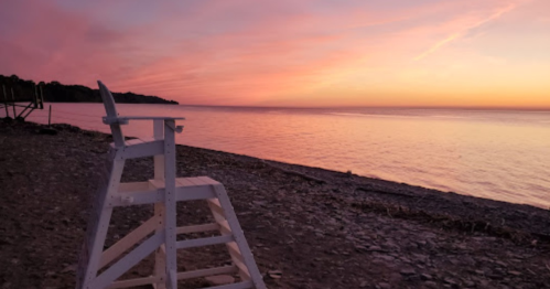 A white lifeguard chair on a rocky beach at sunset, with calm water and colorful sky in the background.