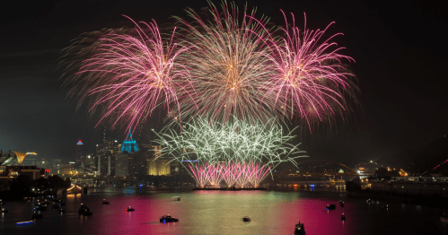 Colorful fireworks burst over a river, illuminating the skyline and boats below in a vibrant night scene.