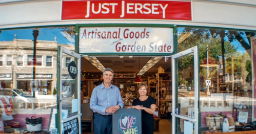 A man and woman stand at the entrance of "Just Jersey," a store featuring artisanal goods, with a welcoming sign.