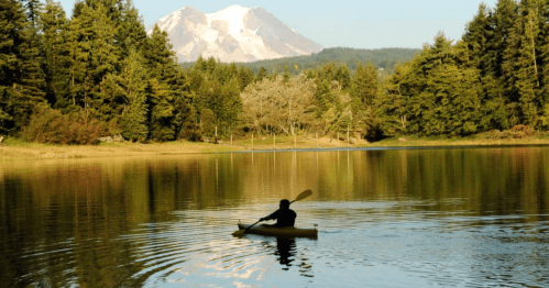 A person kayaking on a calm lake with a snow-capped mountain and lush trees in the background.