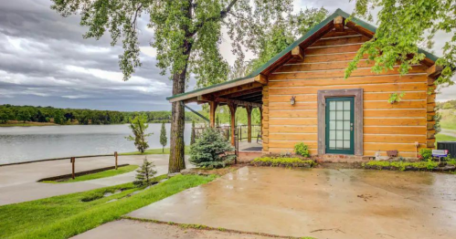 A rustic log cabin by a lake, surrounded by greenery and under a cloudy sky.