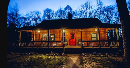 A cozy log cabin at dusk, featuring a red door and warm lights, surrounded by trees.