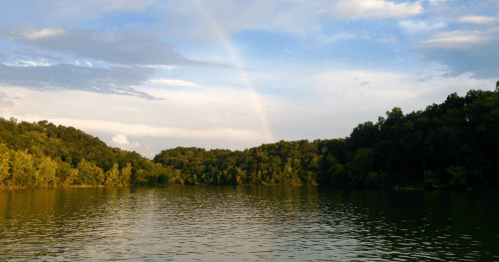 A serene lake surrounded by lush green trees, with a rainbow arching across a partly cloudy sky.