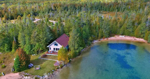 Aerial view of a cozy cabin by a clear blue lake, surrounded by lush green trees and colorful autumn foliage.