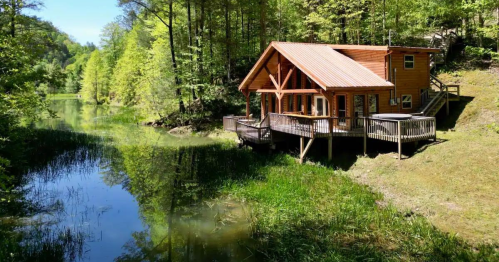 A wooden cabin with a deck by a serene lake, surrounded by lush green trees and reflections in the water.