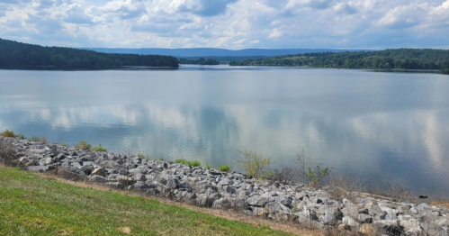 A serene lake surrounded by green hills under a cloudy sky, with a rocky shoreline in the foreground.