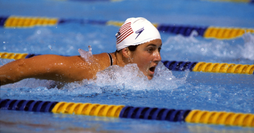 A swimmer in a white cap with an American flag competes in a pool, creating splashes as they perform a stroke.