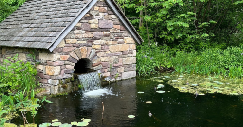 A stone building with a sloped roof beside a pond, featuring a small waterfall and surrounded by lush greenery.