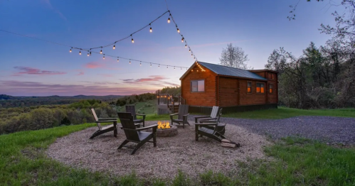 Cozy cabin at sunset with string lights and a fire pit surrounded by chairs in a scenic outdoor setting.