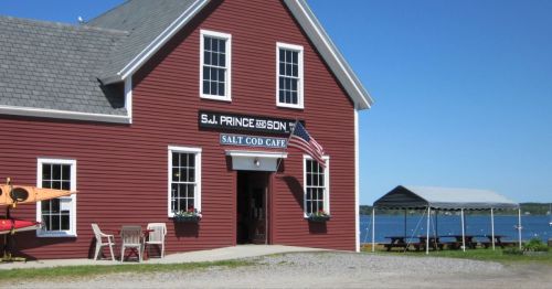 A red building with a sign reading "Salt Cod Cafe," featuring an American flag and a view of the water nearby.