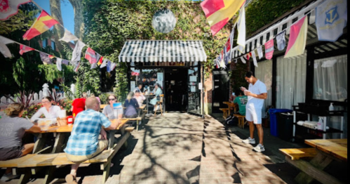 Outdoor dining area with people at picnic tables, colorful flags, and a café entrance surrounded by greenery.