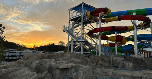 A water park with colorful slides and a tall structure against a sunset sky, surrounded by rocky terrain.