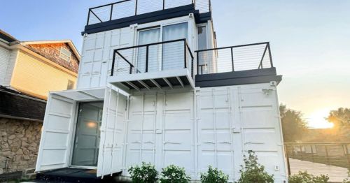 A modern two-story building made from white shipping containers, featuring a balcony and a large entrance.