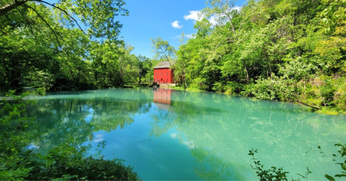 A serene pond surrounded by lush greenery, featuring a red building reflected in the calm, turquoise water.