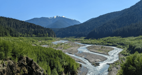 A winding river flows through a lush green valley, surrounded by mountains under a clear blue sky.