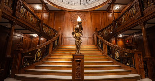 A grand staircase with ornate wooden railings and a statue holding a torch, illuminated by elegant lighting.