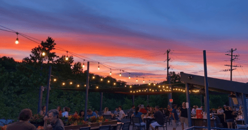 A vibrant sunset over a lively outdoor dining area, with string lights and people enjoying their evening.