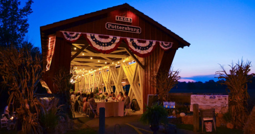 A rustic barn decorated with lights and bunting, set against a twilight sky, with people dining inside.