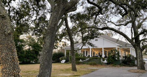 A charming white house surrounded by trees, with outdoor lights and a grassy area in the foreground.