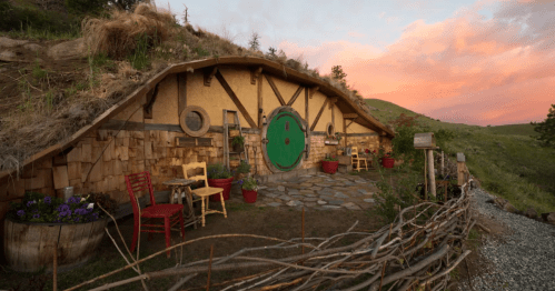 A charming hobbit-style home with a round green door, surrounded by colorful chairs and flower pots at sunset.