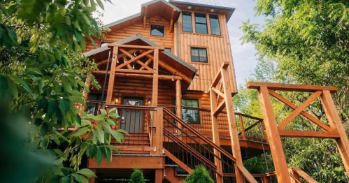 A large wooden cabin with multiple levels, surrounded by greenery and featuring a spacious front porch and stairs.