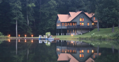 A serene lakeside house with a wooden deck, surrounded by trees, reflecting in calm water at dusk.