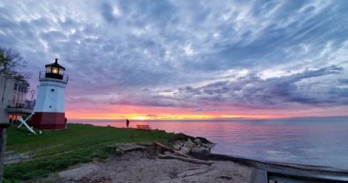 A lighthouse by the shore at sunset, with vibrant clouds and calm waters reflecting the colorful sky.