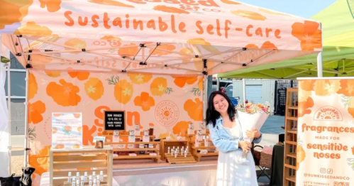 A woman stands at a colorful market stall promoting sustainable self-care products, holding a bouquet of flowers.