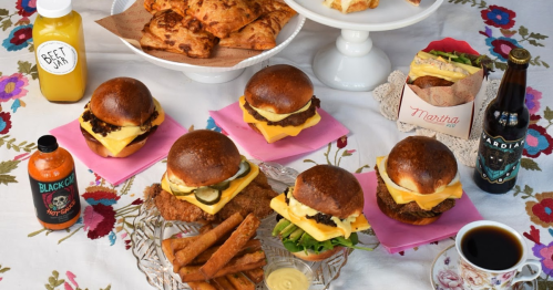A colorful spread of burgers, snacks, drinks, and pastries on a floral tablecloth.