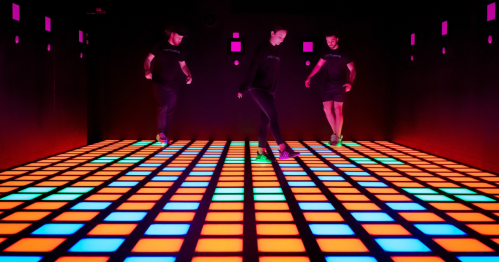 Three people dance on a colorful, illuminated floor with vibrant squares in a dark room.