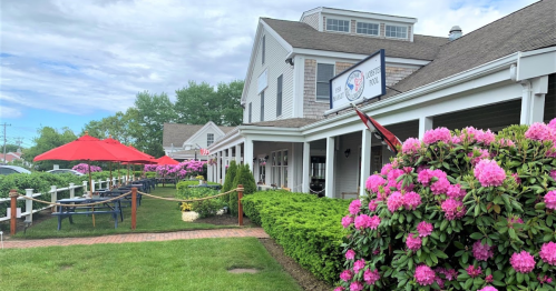 A charming restaurant exterior with pink flowers, green bushes, and red umbrellas on a patio under a cloudy sky.