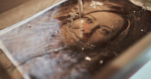 A close-up of a woman's face partially submerged in water, with water flowing over the image.