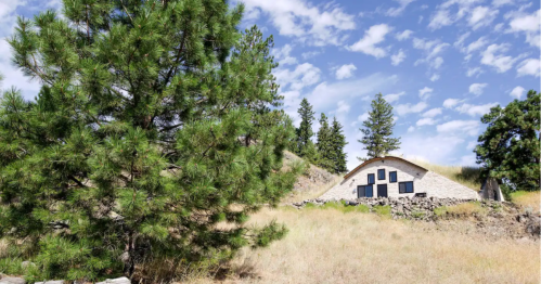 A rustic house nestled among tall trees and grassy hills under a blue sky with scattered clouds.