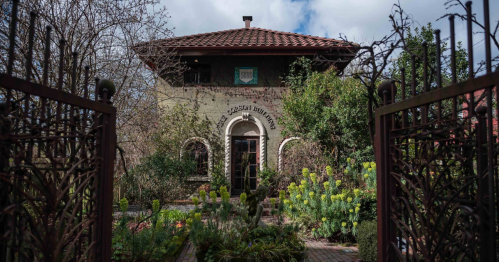 A charming house with a tiled roof, surrounded by lush greenery and flowers, viewed through ornate metal gates.