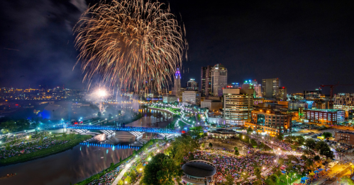 A vibrant city skyline illuminated by colorful fireworks over a river, with a crowd gathered below.