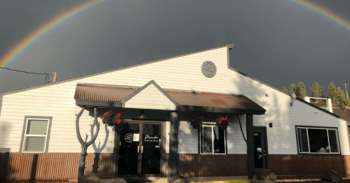 A white building with a metal roof, featuring flower baskets, under a rainbow against a dark sky.