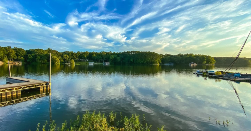 A serene lake scene with calm water, lush greenery, and a blue sky filled with clouds, reflecting on the surface.