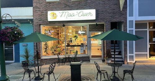 A cozy storefront with a sign reading "Moo-Chew," featuring outdoor seating and green umbrellas.