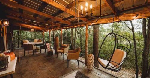 Cozy outdoor porch with hanging chair, seating area, and rustic decor, surrounded by trees. Warm, inviting atmosphere.