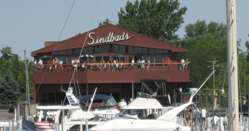 A marina with boats in the foreground and a restaurant named "Sindbads" bustling with people on the balcony.