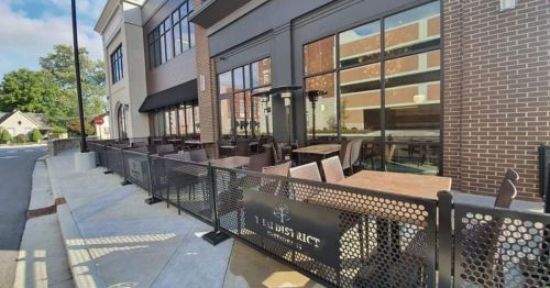 Outdoor dining area of a restaurant with tables and chairs, surrounded by a black fence and large windows.