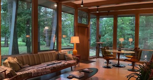 A cozy living room with large windows, a sofa, a round dining table, and a lamp, surrounded by greenery.
