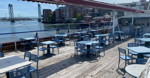 Outdoor dining area on a boat, featuring blue tables and chairs, with a view of the water and nearby buildings.