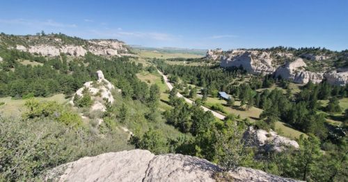 A panoramic view of a lush green valley surrounded by rocky cliffs and a winding road under a clear blue sky.