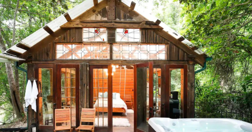 Cozy wooden cabin with large glass doors, surrounded by greenery, featuring a hot tub and outdoor seating.