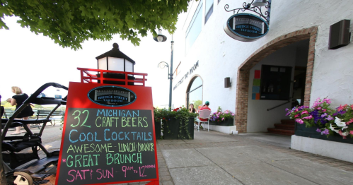 A sign outside a restaurant advertises craft beers, cocktails, and brunch hours, surrounded by flowers and outdoor seating.