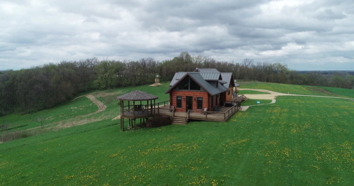 A rustic wooden house on a grassy hill, surrounded by trees and a cloudy sky, with a deck and gazebo nearby.