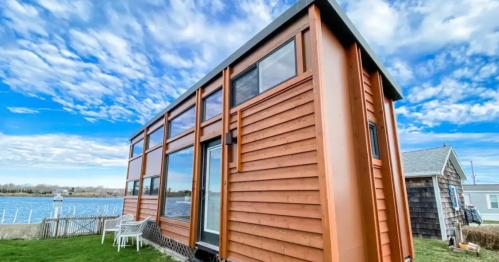 A modern tiny house with wooden siding, large windows, and a scenic view of water and clouds in the background.