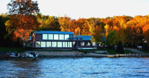 A modern lakeside house surrounded by vibrant autumn foliage and calm water.